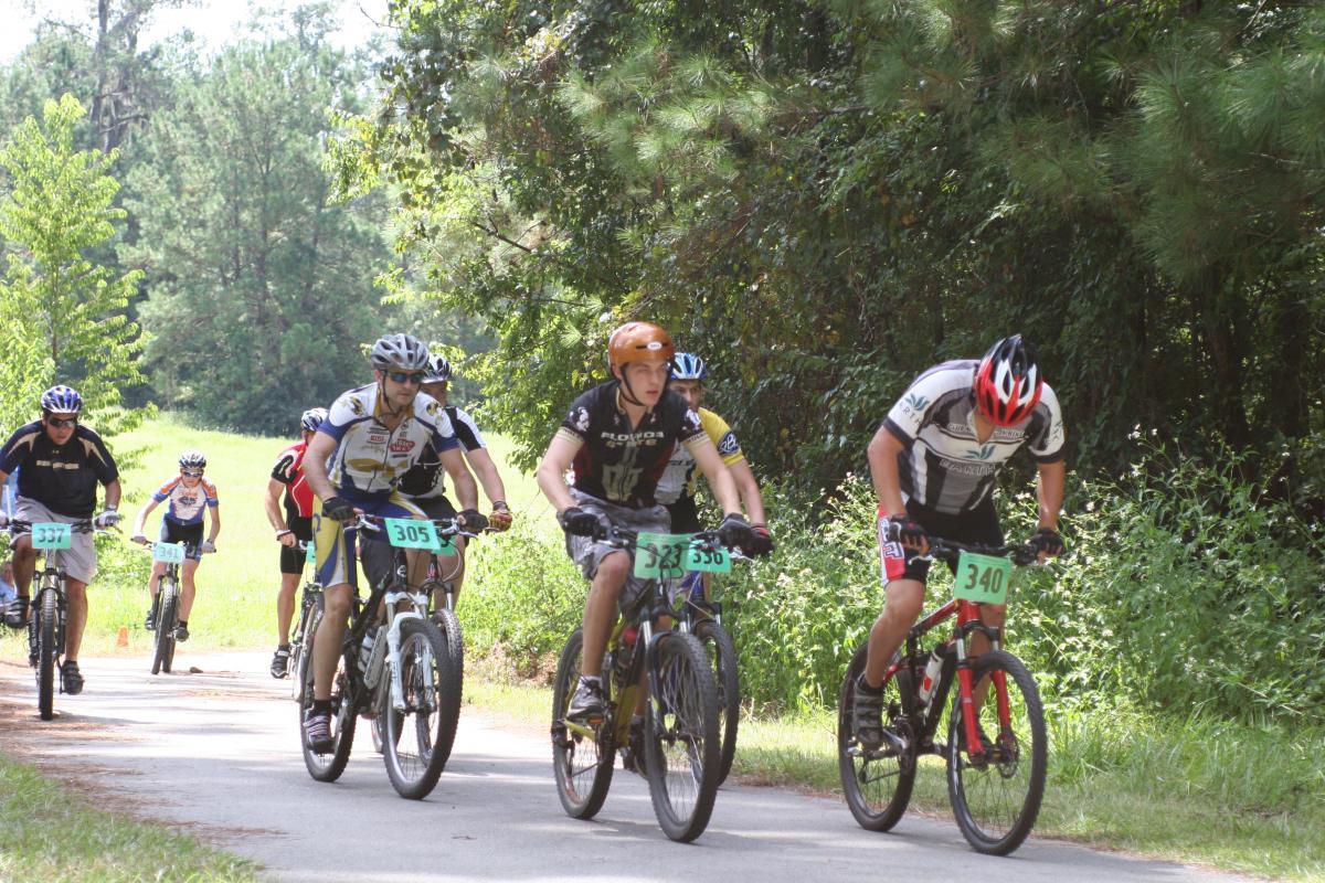 A group of five male mountain bikers riding on a narrow path surrounded by trees and grass. They wear helmets and cycling gear, each displaying a number on their bikes. The scene captures the action of biking in a natural outdoor setting, with bright sunlight filtering through the foliage. The Rock Trail mountain bike trail.