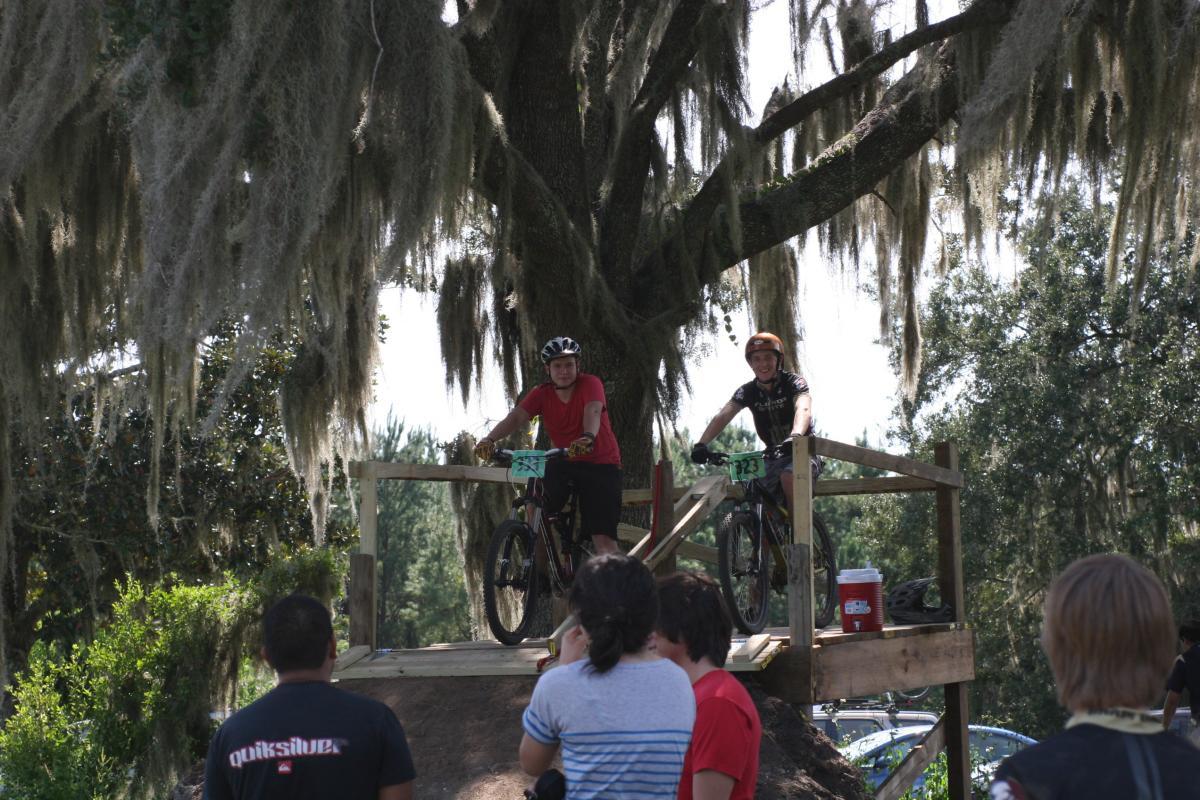 Two mountain bikers are positioned on a wooden platform built into the side of a dirt ramp, preparing to ride down. One rider in a red shirt and the other in a black shirt with the number 23 displayed on their bike are smiling and focused. In the foreground, several spectators are watching, while large trees draped with Spanish moss create a natural backdrop. The Rock Trail mountain bike trail.