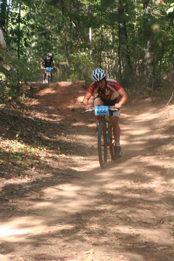 A mountain bike racer, wearing a red jersey and white shorts with the number 222, rides along a dirt trail surrounded by trees. In the background, another cyclist is also navigating the path, creating a sense of action in the forested area. Issaqueena Lake mountain bike trail.