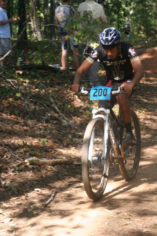 A mountain biker competes in a race on a wooded trail, wearing a black jersey and blue helmet, with the number 200 displayed on his bike. The terrain is covered in dirt and fallen leaves, and other cyclists can be seen in the background. Issaqueena Lake mountain bike trail.