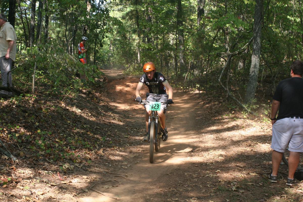 A mountain bike racer in an orange helmet and a black jersey with the number 23 rides down a dirt trail surrounded by trees. Two spectators watch from the side, one wearing light-colored shorts and a black shirt, and the other in a red cycling outfit. Dust rises from the path as the racer navigates the course. Issaqueena Lake mountain bike trail.