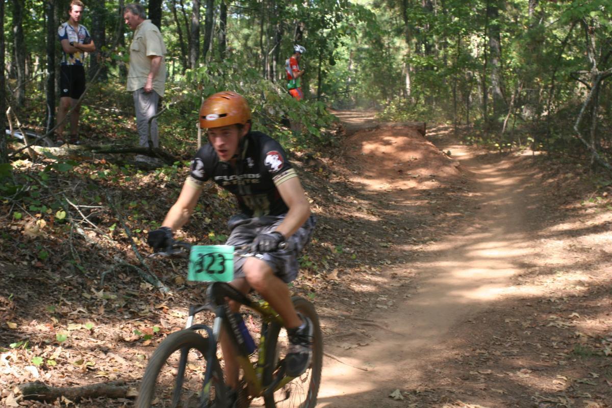 A mountain biker in motion, wearing a black jersey and an orange helmet, rides along a dirt trail in a wooded area. The biker has a race number pinned to their front. In the background, two spectators observe the race, surrounded by trees and fallen leaves. Issaqueena Lake mountain bike trail.