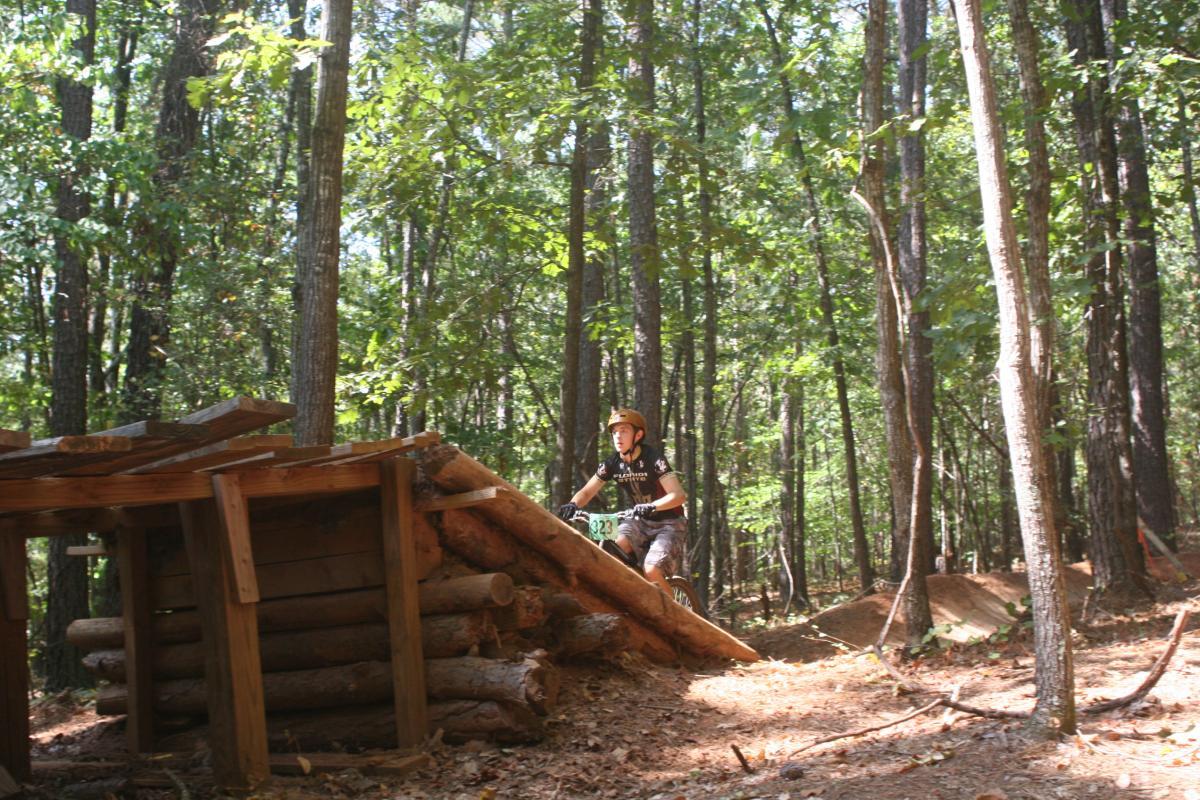 A cyclist navigating a wooden ramp in a forested area, surrounded by tall trees and dappled sunlight. The cyclist is wearing a helmet and riding a mountain bike, while the ramp is constructed from logs and wooden planks. Issaqueena Lake mountain bike trail.