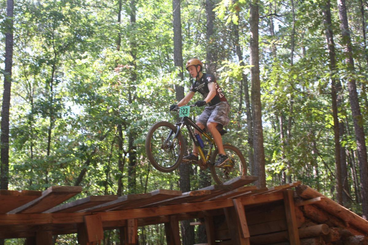 A young cyclist with a helmet and racing number 223 performs a jump on a mountain bike, navigating a wooden ramp in a wooded area, surrounded by trees and sunlight. Issaqueena Lake mountain bike trail.