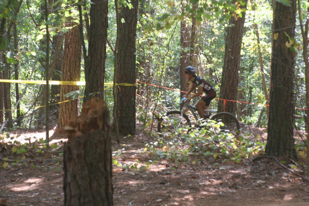 A mountain biker navigating a wooded trail, surrounded by tall trees and caution tape, with sunlight filtering through the leaves. Issaqueena Lake mountain bike trail.