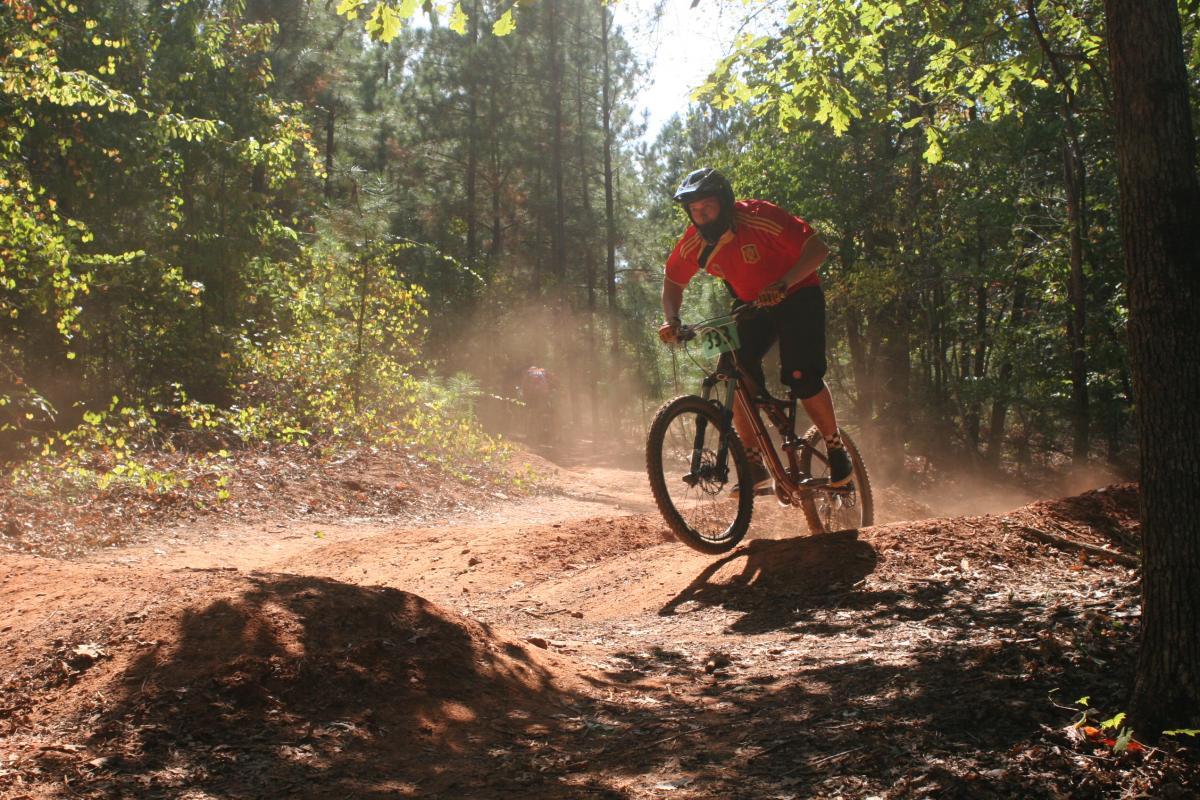 A mountain biker in a red jersey navigates a dirt trail, capturing air as he jumps off a small mound. Surrounding him are trees and a cloud of dust, indicating a lively outdoor biking environment. Sunlight filters through the leaves, creating a vibrant atmosphere. Issaqueena Lake mountain bike trail.