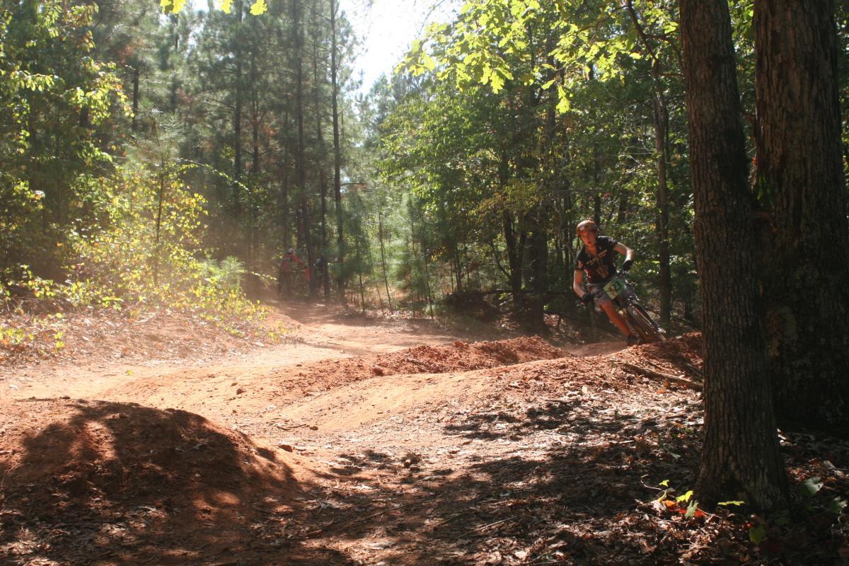 A cyclist navigating a dirt trail in a wooded area, kicking up dust as they ride around a bend. Sunlight filters through the trees, illuminating the path and surrounding greenery. Issaqueena Lake mountain bike trail.