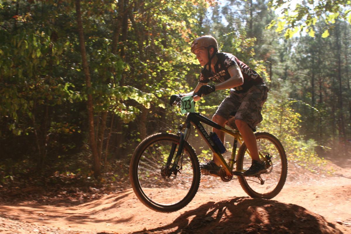 A mountain biker in mid-air, leaping off a dirt jump on a forest trail. The rider wears a helmet and a jersey with a race number, and is surrounded by trees and dust in the sunlight. Issaqueena Lake mountain bike trail.