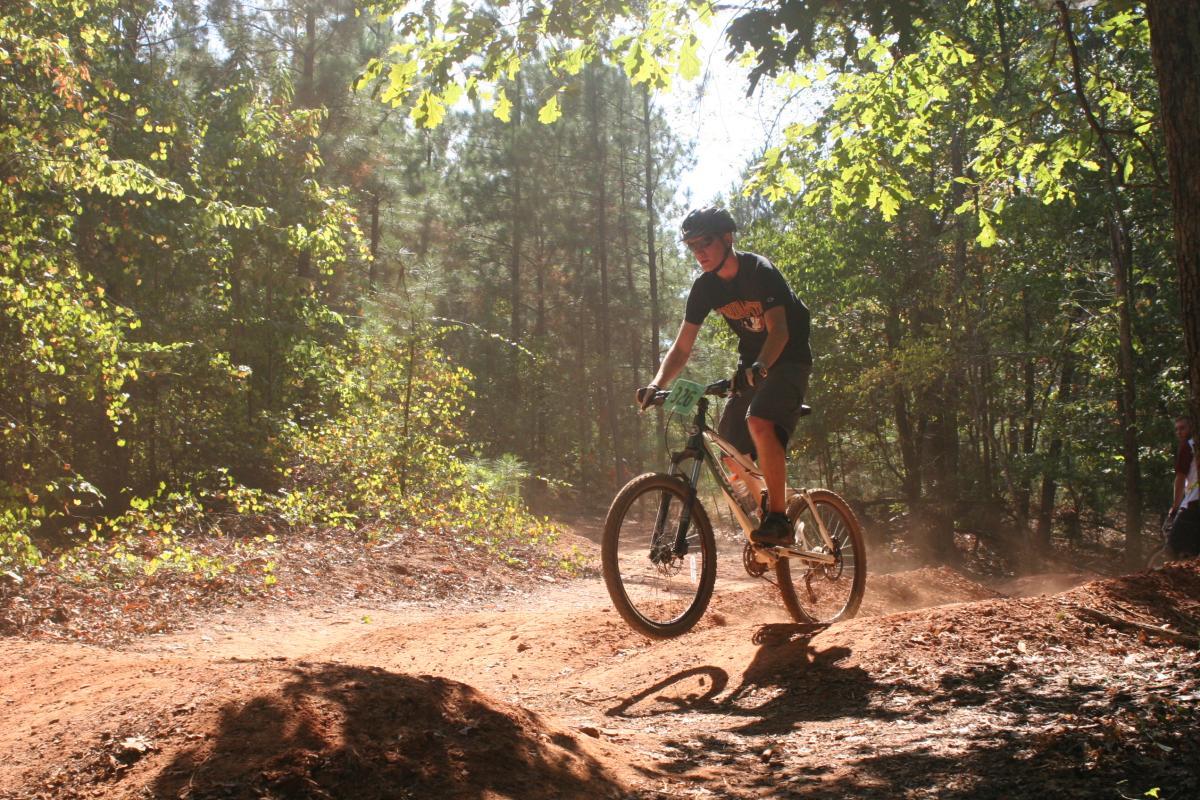 A mountain biker in a black shirt and helmet rides along a dirt trail in a forest, surrounded by trees with sunlight filtering through the leaves. Dust is kicked up as the rider approaches a small jump, showcasing an active outdoor recreation scene. Issaqueena Lake mountain bike trail.