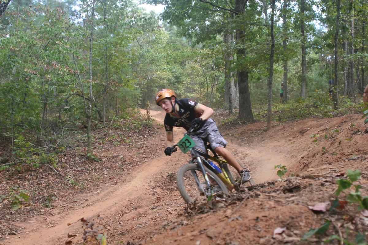 A young cyclist wearing a helmet and gloves leans into a turn on a dirt mountain biking trail, surrounded by trees and foliage. The cyclist's bike is outfitted for off-road riding, and a green race number is visible on their shirt. Dust is being kicked up from the trail as they navigate the winding path. Issaqueena Lake mountain bike trail.