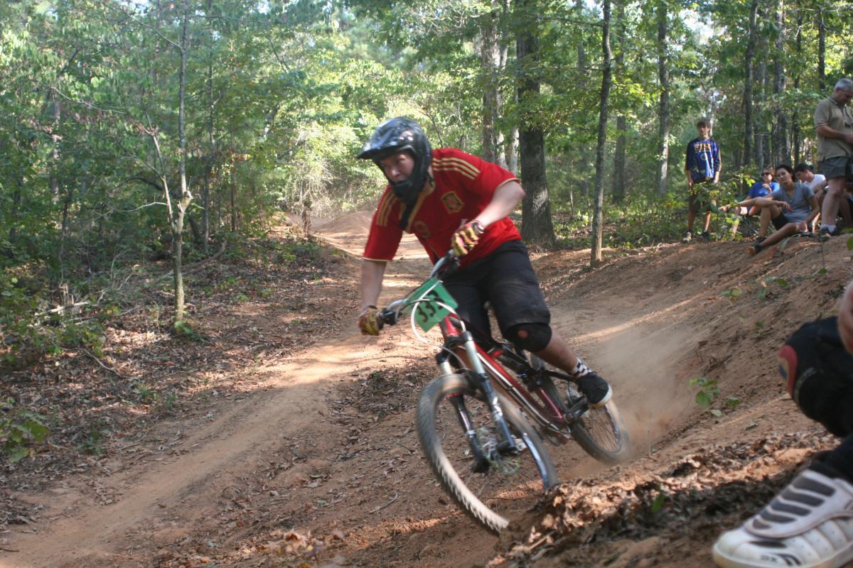 A mountain biker in a red shirt navigates a dirt trail surrounded by trees. The rider leans into a curve, kicking up dust as they accelerate, while several onlookers watch from the side. Issaqueena Lake mountain bike trail.