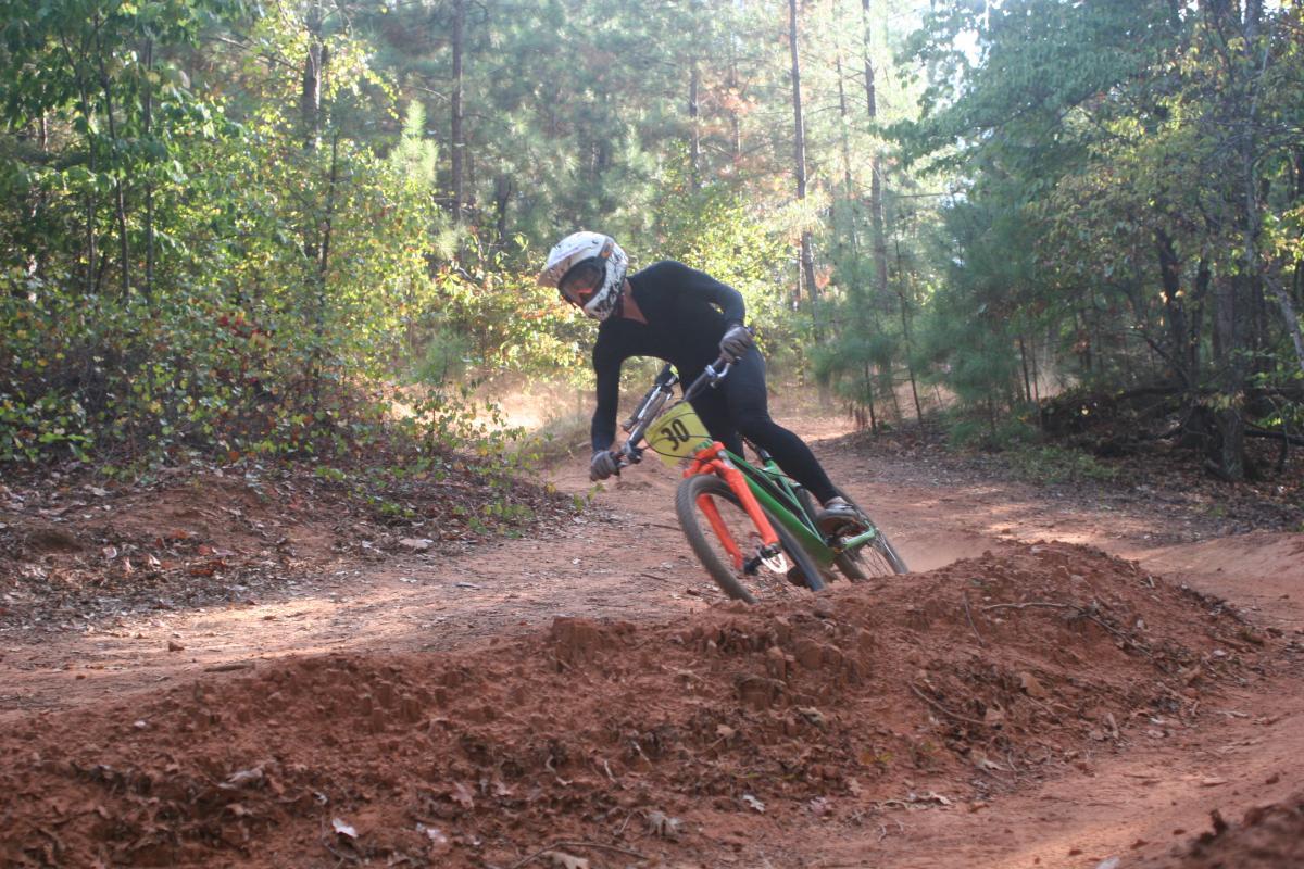 A cyclist in a black outfit rides a mountain bike on a dirt trail in a wooded area. The bike features an orange front wheel and has a race number attached. The cyclist leans into a turn while navigating over a dirt jump, surrounded by trees and greenery. Issaqueena Lake mountain bike trail.