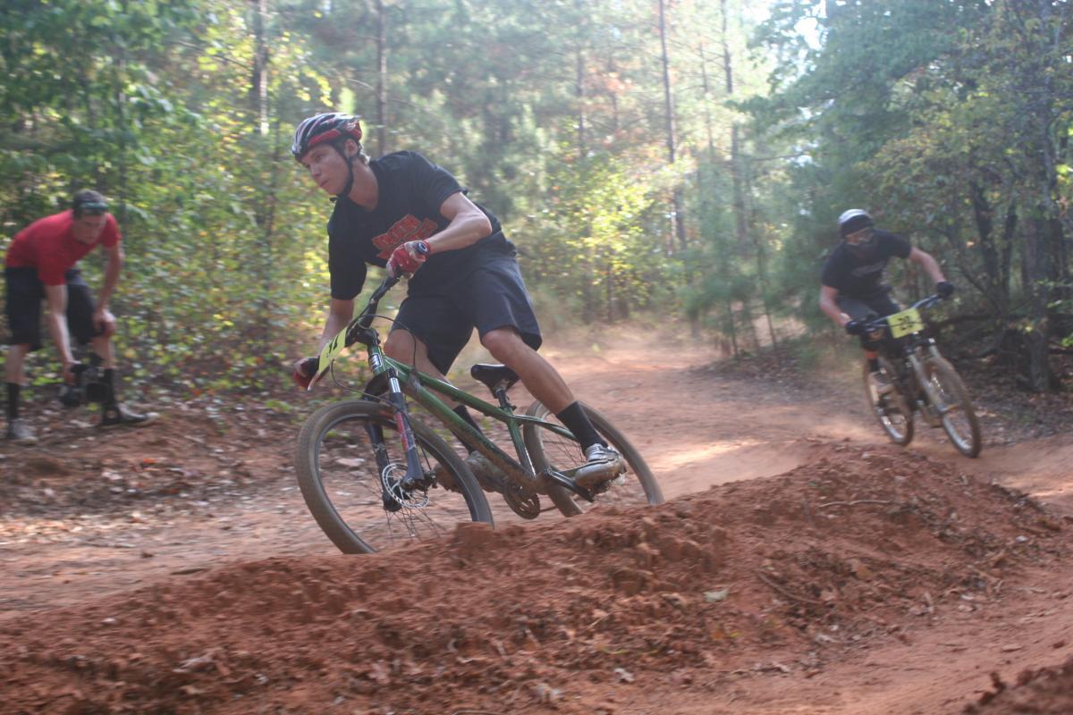 Two mountain bikers navigating a dirt trail in a forested area. One rider, wearing a black shirt and helmet, is leaning into a turn on a green bicycle, while another rider in the background is approaching the scene. The ground is covered in reddish dirt, and the background features trees and foliage, suggesting an outdoor biking event. Issaqueena Lake mountain bike trail.
