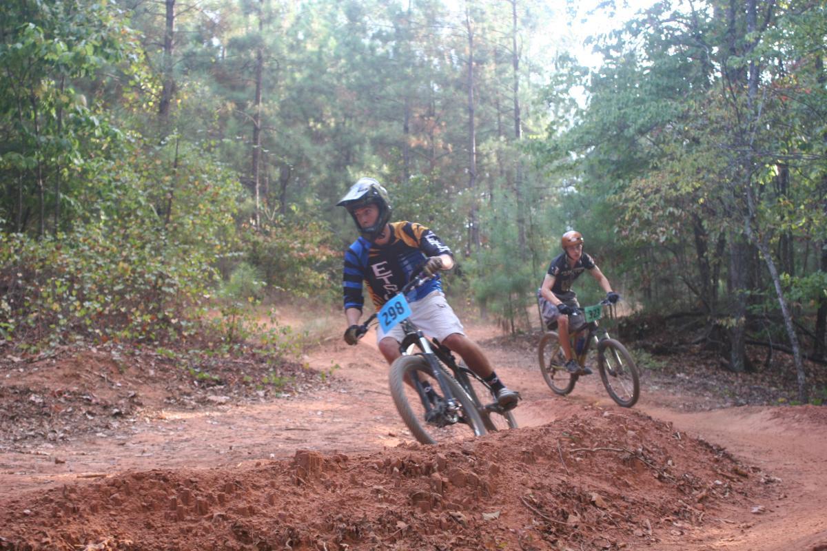 Two mountain bikers navigate a dirt trail in a forested area, with trees and green foliage in the background. One rider, wearing a blue and black jersey marked with the number 298, leans into a turn while the other, in a black jersey marked with the number 32, follows closely behind. The ground is rough, with a dirt mound visible nearby. Issaqueena Lake mountain bike trail.