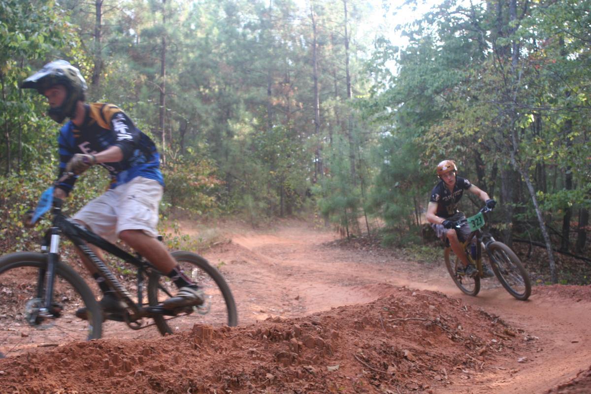 Two mountain bikers navigating a dirt trail in a wooded area. One cyclist, wearing a blue and black jersey, is slightly blurred, indicating motion as he rides past a dirt jump. The second cyclist, wearing a black jersey with the number 323 and a helmet, leans into the turn on the trail. The surrounding landscape features trees with green foliage, creating a natural, outdoor setting. Issaqueena Lake mountain bike trail.