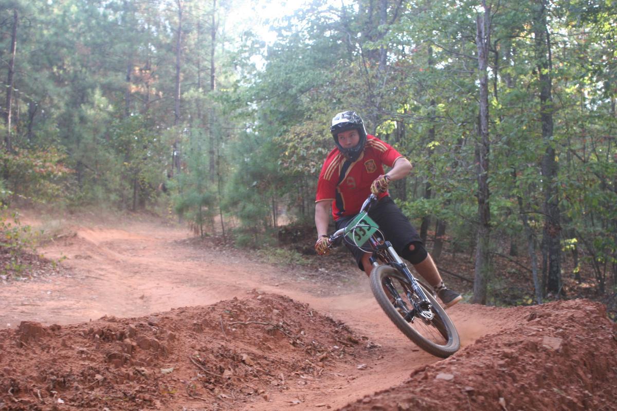 A mountain biker in a red jersey navigates a dirt trail in a forested area, leaning into a turn on a dirt jump. The surrounding trees are lush and green, and dust is kicked up from the trail as the biker rides. Issaqueena Lake mountain bike trail.
