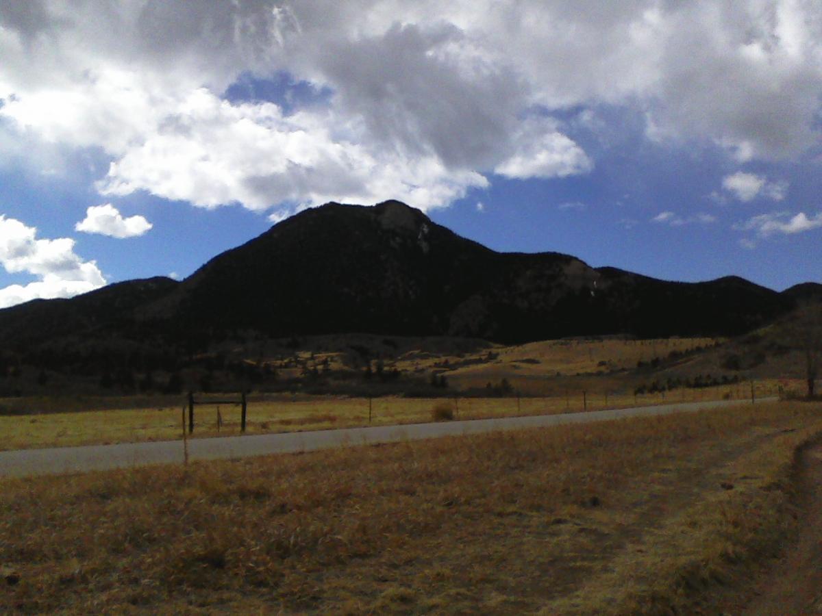 A scenic view of a mountain against a partly cloudy sky, with a field in the foreground and a winding road. The landscape features golden grass and sparse trees, creating a tranquil outdoor setting. Falcon Trail mountain bike trail.
