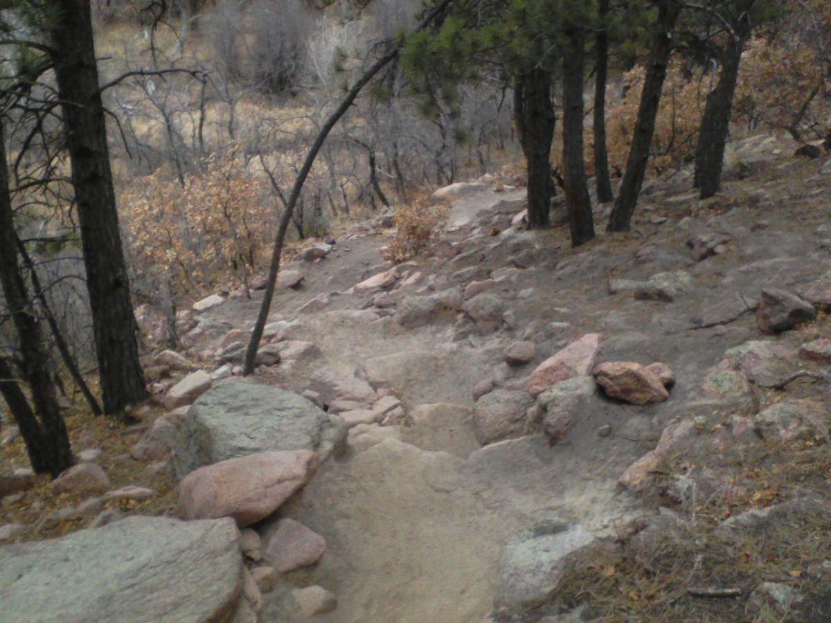 A narrow dirt path winding through a rocky landscape, flanked by trees and dry, autumn foliage. The ground is uneven with large boulders and scattered pebbles, creating a natural hiking trail. Falcon Trail mountain bike trail.