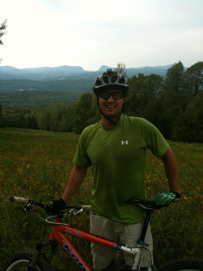 A smiling cyclist wearing a green shirt and a helmet stands beside an orange mountain bike on a grassy slope, with a scenic view of mountains and a forest in the background. The sky is overcast, suggesting an outdoor ride in nature. Kingdom Trails mountain bike trail.