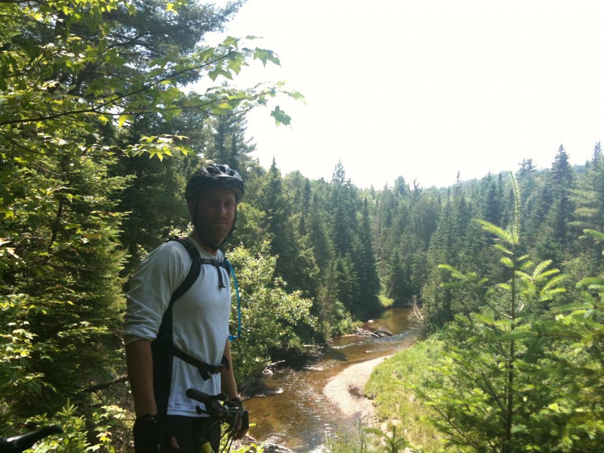 A cyclist wearing a helmet and a light-colored long-sleeve shirt stands near a winding river, surrounded by lush green trees and foliage. The sunlight filters through the leaves, creating a peaceful and scenic outdoor setting. Kingdom Trails mountain bike trail.