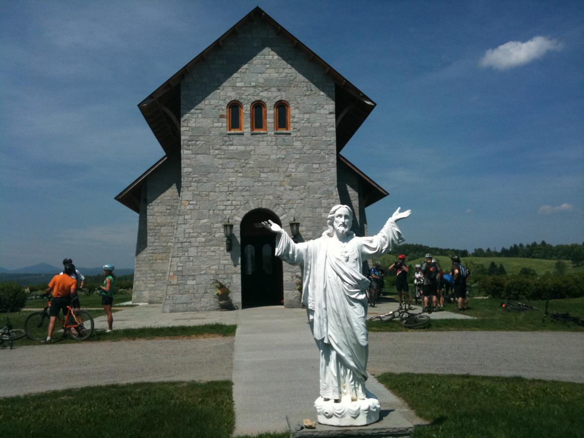 A stone church with a triangular roof stands against a blue sky. In the foreground, a white statue of Jesus with outstretched arms is positioned on the pathway. Cyclists in casual attire are gathered nearby, some with bicycles, enjoying the scenic view of green rolling hills in the background. Kingdom Trails mountain bike trail.