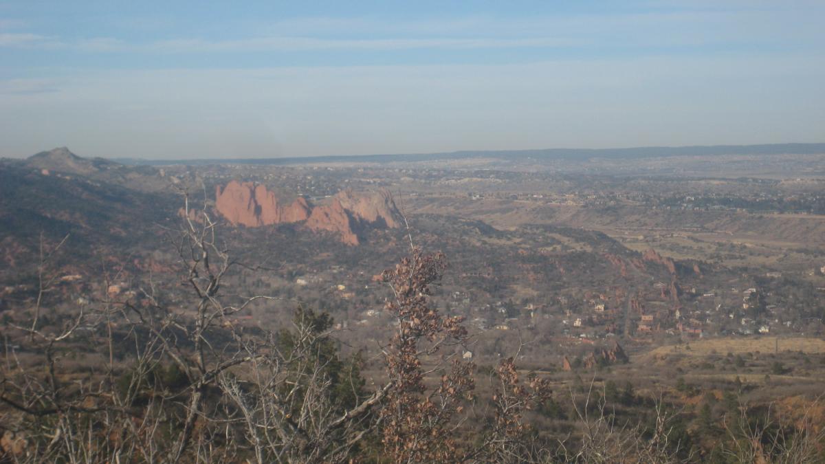 A panoramic view of a mountainous landscape featuring red rock formations in the distance, with a valley and small towns visible below. The sky is clear with soft clouds, and bushes with brown leaves are in the foreground. Paul Intemann Memorial Trail mountain bike trail.