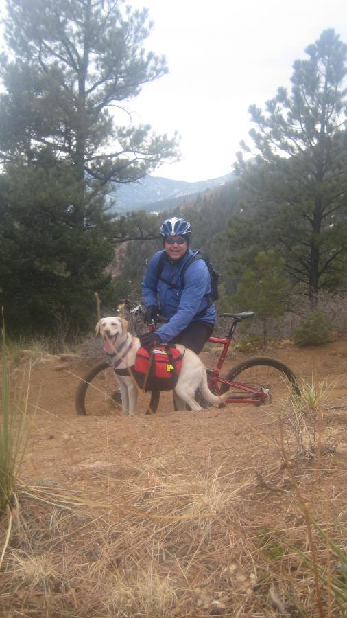 A person wearing a blue jacket and helmet sits on a mountain bike next to a dog with a backpack. They are surrounded by trees and a rocky terrain, with distant mountains visible in the background. The scene captures a sense of outdoor adventure and companionship. Captain Jack's mountain bike trail.