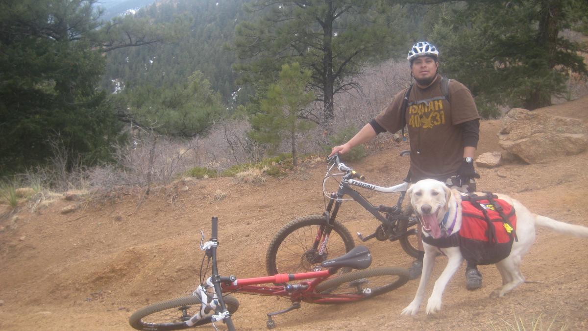 A person wearing a helmet and casual clothing stands next to a mountain bike on a dirt path, with a friendly dog equipped with a backpack nearby. The background features trees and hills, suggesting a scenic outdoor location ideal for biking and hiking. Captain Jack's mountain bike trail.