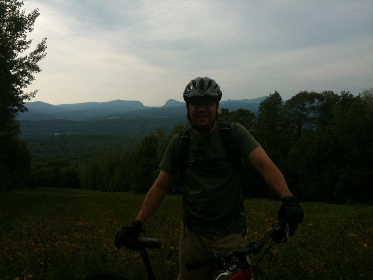 A person wearing a helmet and gloves stands next to a mountain bike on a grassy slope, with a scenic view of distant mountains and a forested valley in the background. The sky is partly cloudy, creating a serene outdoor atmosphere. Kingdom Trails mountain bike trail.