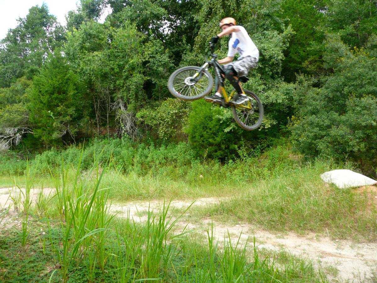 A person riding a mountain bike performs a jump above a dirt path, surrounded by lush green vegetation. The cyclist is wearing a white shirt and patterned shorts, with a cap on their head, showcasing an action-packed moment in a natural outdoor setting. Santos mountain bike trail.