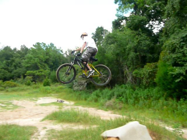 A person wearing a helmet and a white shirt is mid-air while performing a jump on a mountain bike over a dirt ramp. The surrounding area features lush green trees and grassy terrain, emphasizing an outdoor biking environment. Santos mountain bike trail.