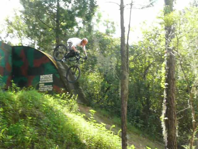 A mountain biker performing a jump off a curved ramp in a wooded area, surrounded by trees and greenery. The biker is wearing a white shirt and is mid-air above the ramp, showcasing an action-packed moment in the sport. Santos mountain bike trail.