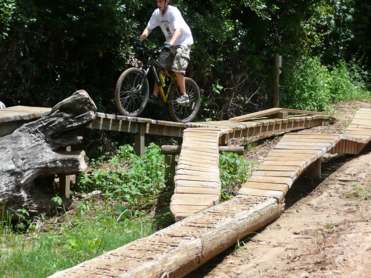A person riding a mountain bike on a wooden trail that includes curved and elevated sections. Surrounding greenery and a log are visible on the side of the trail, suggesting a natural outdoor setting for biking. Tom Brown / Lafayette Heritage Park mountain bike trail.