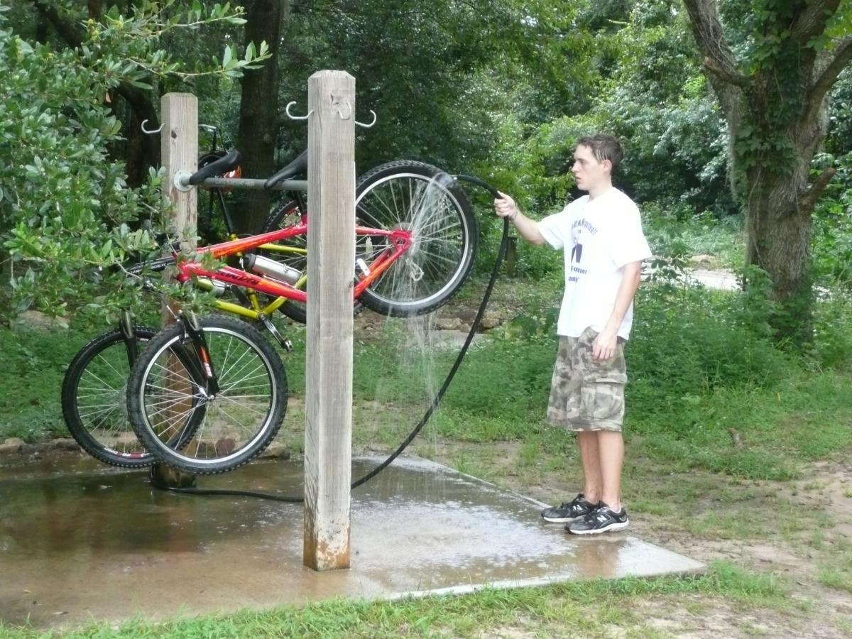 A young person using a hose to wash a bike mounted on a bike rack in a green outdoor setting. Water sprays from the hose, creating a small puddle on the ground. The background features trees and vegetation. Tom Brown / Lafayette Heritage Park mountain bike trail.