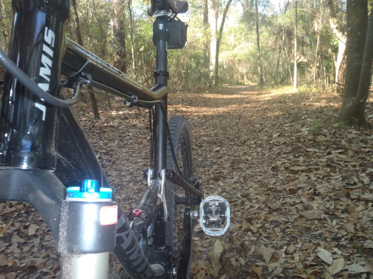 A close-up view of a black mountain bike in a forested area, with a dirt trail covered in fallen leaves in the background. The bike is angled to show the frame and pedal, with sunlight filtering through the trees. Chacala Trail mountain bike trail.