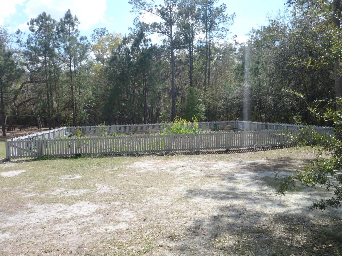 A circular garden enclosed by a white picket fence, surrounded by trees, with a gravel and grass area in the foreground. The garden has a few green plants visible inside the fence, and the sky is partly cloudy. The scene is peaceful and rural. Creekside mountain bike trail.