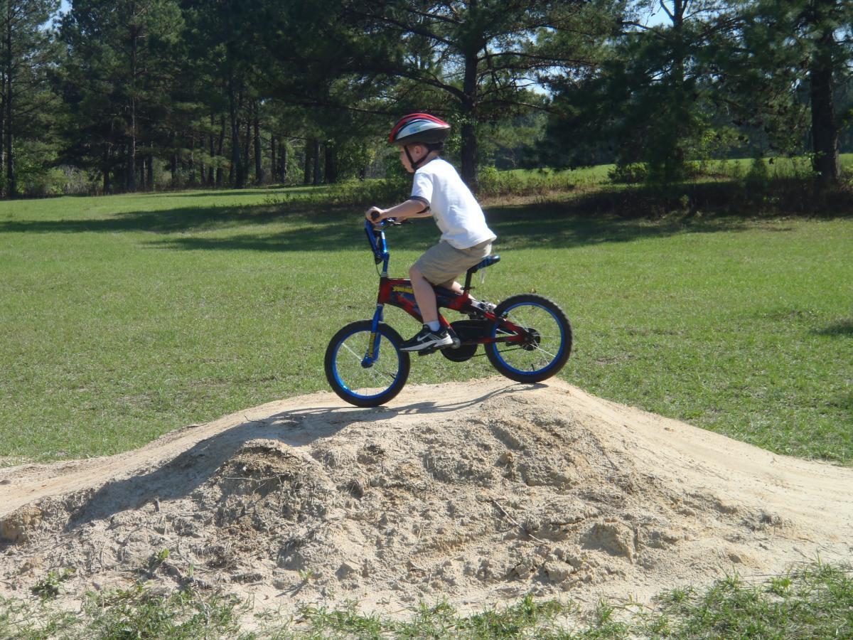 A young boy wearing a helmet rides a small bicycle on top of a dirt mound in a grassy area, surrounded by trees. The sun is shining, indicating a clear day. The Rock Trail mountain bike trail.