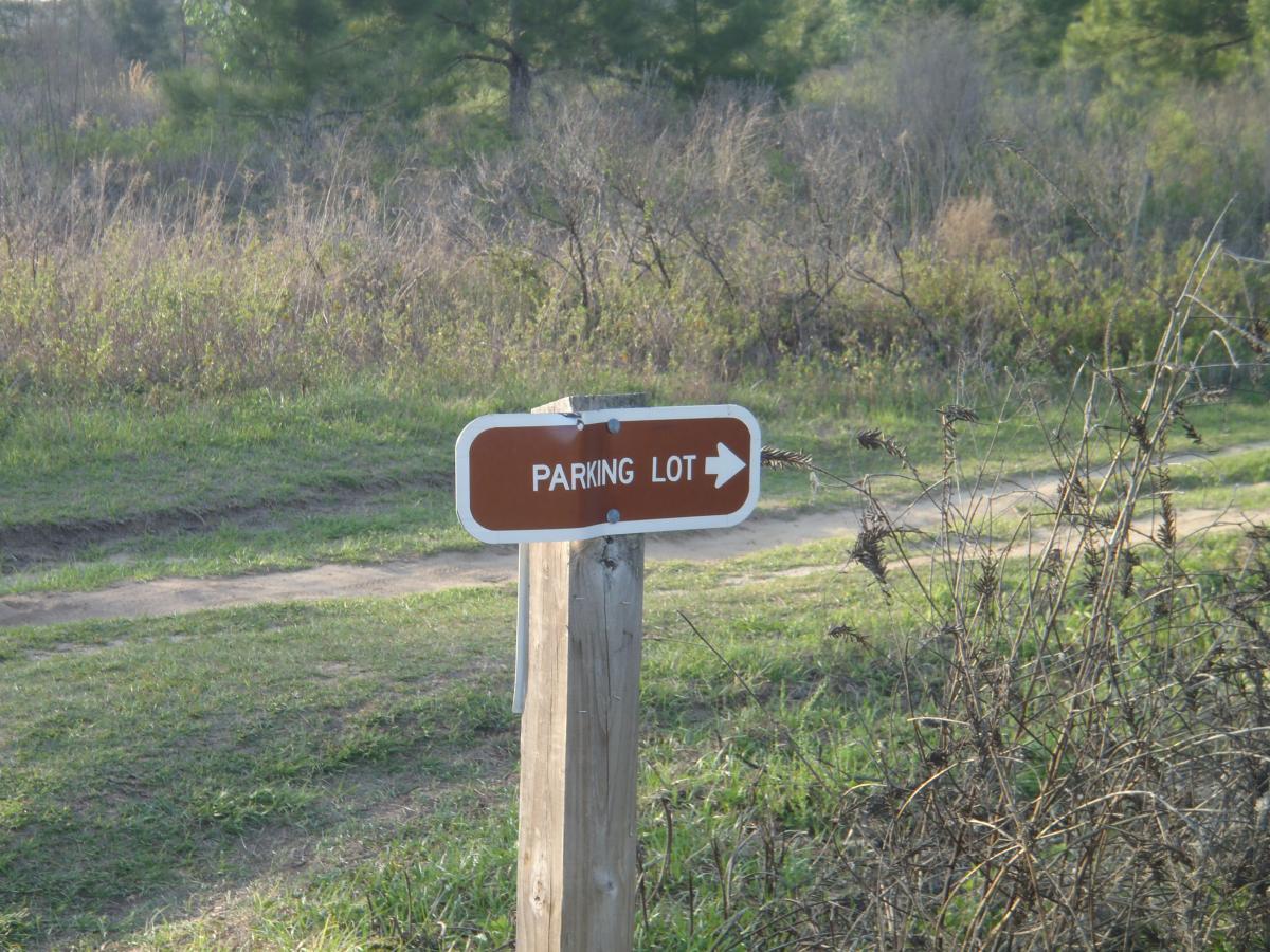 A sign indicating the direction to a parking lot, featuring brown lettering on a white background, mounted on a wooden post in a grassy area with sparse vegetation. San Felasco Hammock Preserve mountain bike trail.
