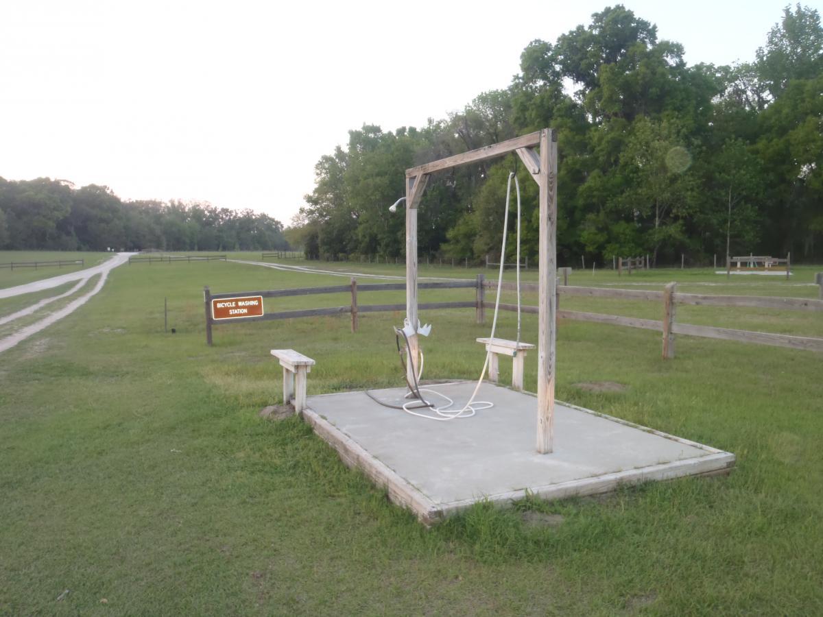 Bicycle washing station with a wooden frame and hose, surrounded by grass and pathways in a park setting. A bench is positioned nearby, with a sign indicating the station's purpose. Lush trees are visible in the background. San Felasco Hammock Preserve mountain bike trail.
