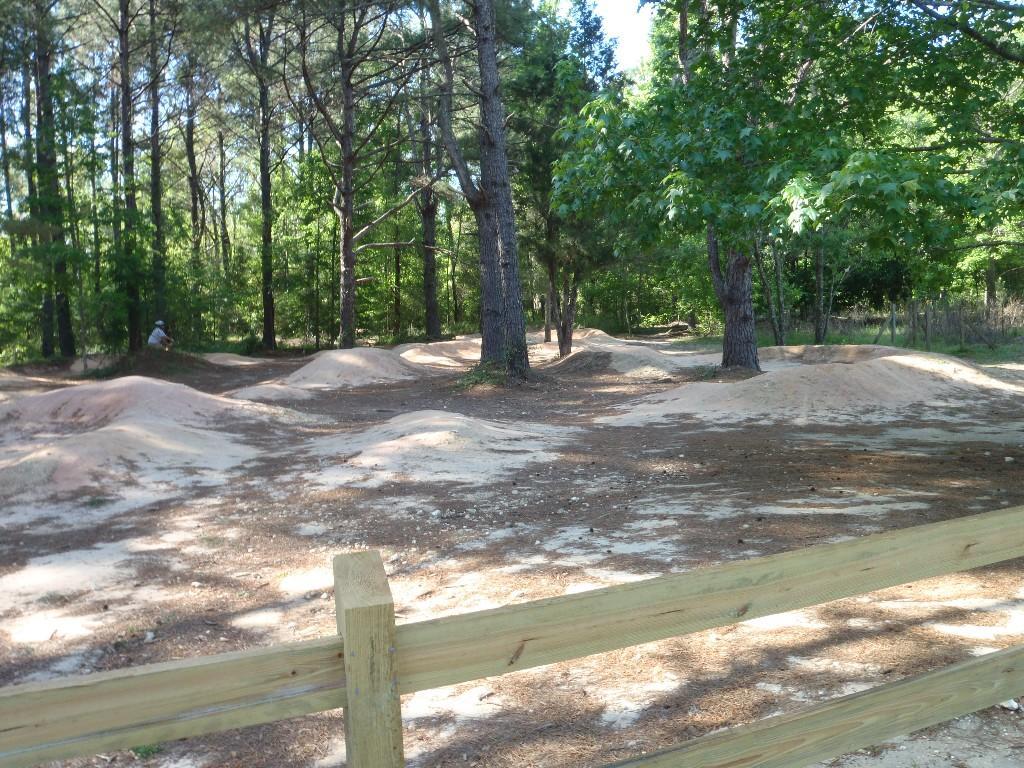 Dirt biking trail with several small dirt jumps surrounded by trees, with a wooden fence in the foreground. Santos mountain bike trail.