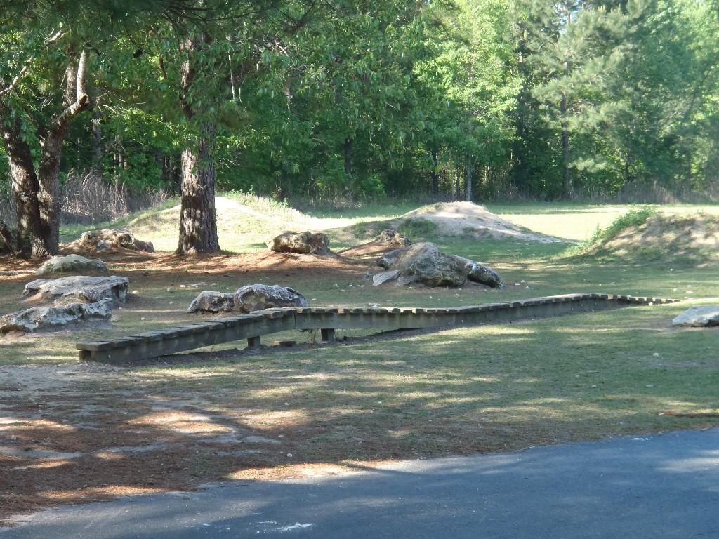 A grassy area featuring a winding wooden walkway surrounded by large rocks and trees. The scene is bright and sunny, with dappled sunlight filtering through the leaves. In the background, there are gentle hills and more trees, creating a natural and serene outdoor setting. Santos mountain bike trail.