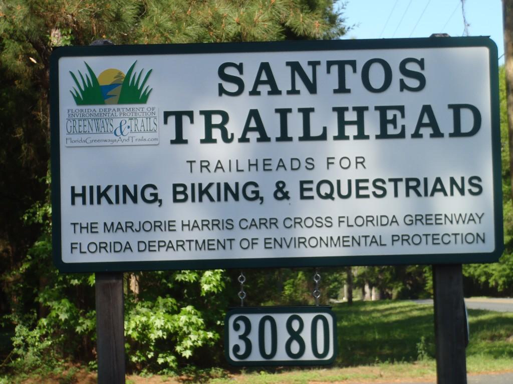 Sign for Santos Trailhead, indicating trail access for hiking, biking, and equestrians. Includes information about the Marjorie Harris Carr Cross Florida Greenway and the Florida Department of Environmental Protection. The sign is located in a wooded area, displaying the number 3080. Santos mountain bike trail.