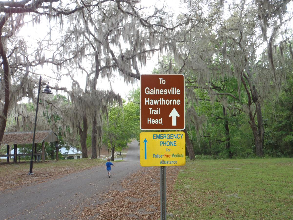A brown directional sign indicating the way to the Gainesville-Hawthorne Trail Head, with an emergency phone sign below it. The background features trees draped with Spanish moss, and a path leading into the distance where a child can be seen running. Gainesville-Hawthorne State Trail mountain bike trail.