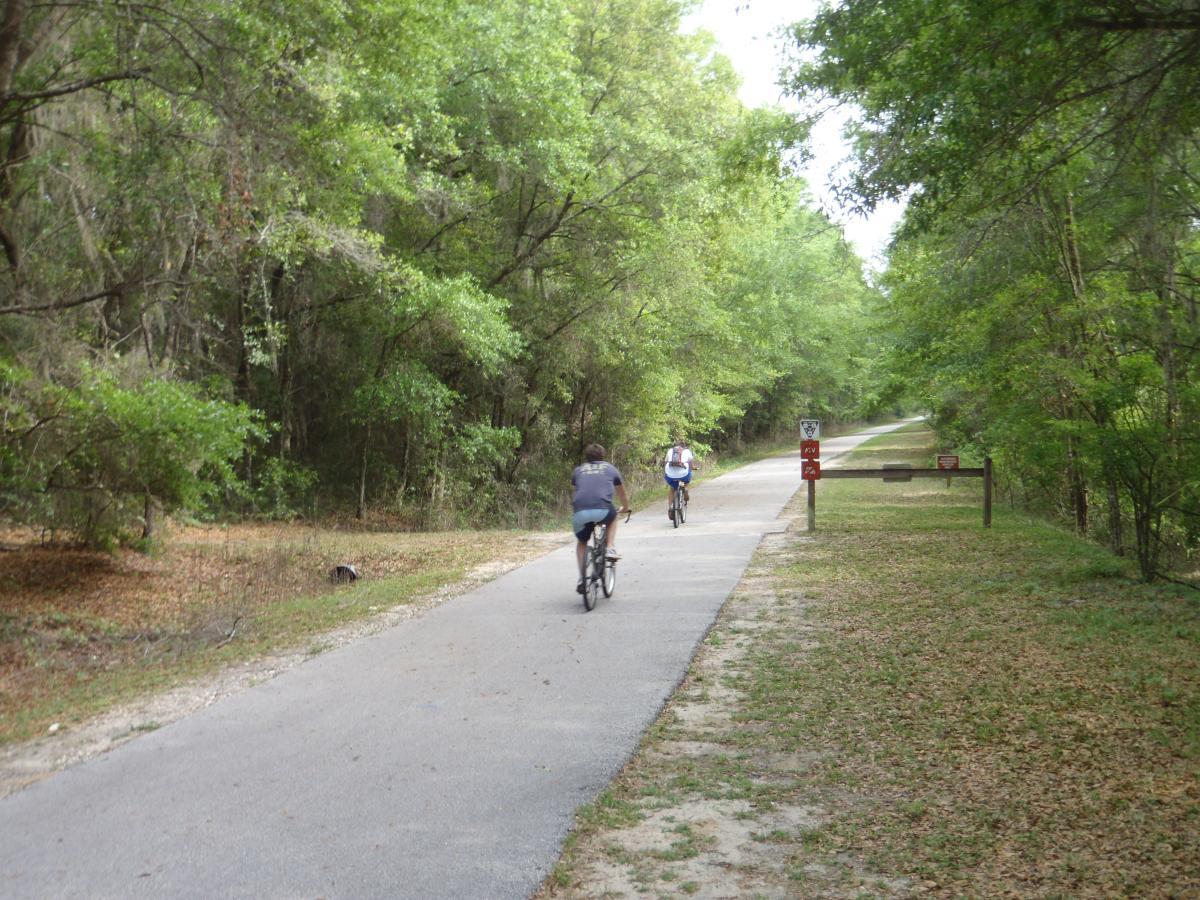 Two cyclists riding on a paved trail surrounded by lush green trees and foliage, with a clear path ahead. A signpost is visible in the background, indicating the recreational area. The ground is covered with scattered leaves, suggesting a natural and serene outdoor setting. Gainesville-Hawthorne State Trail mountain bike trail.