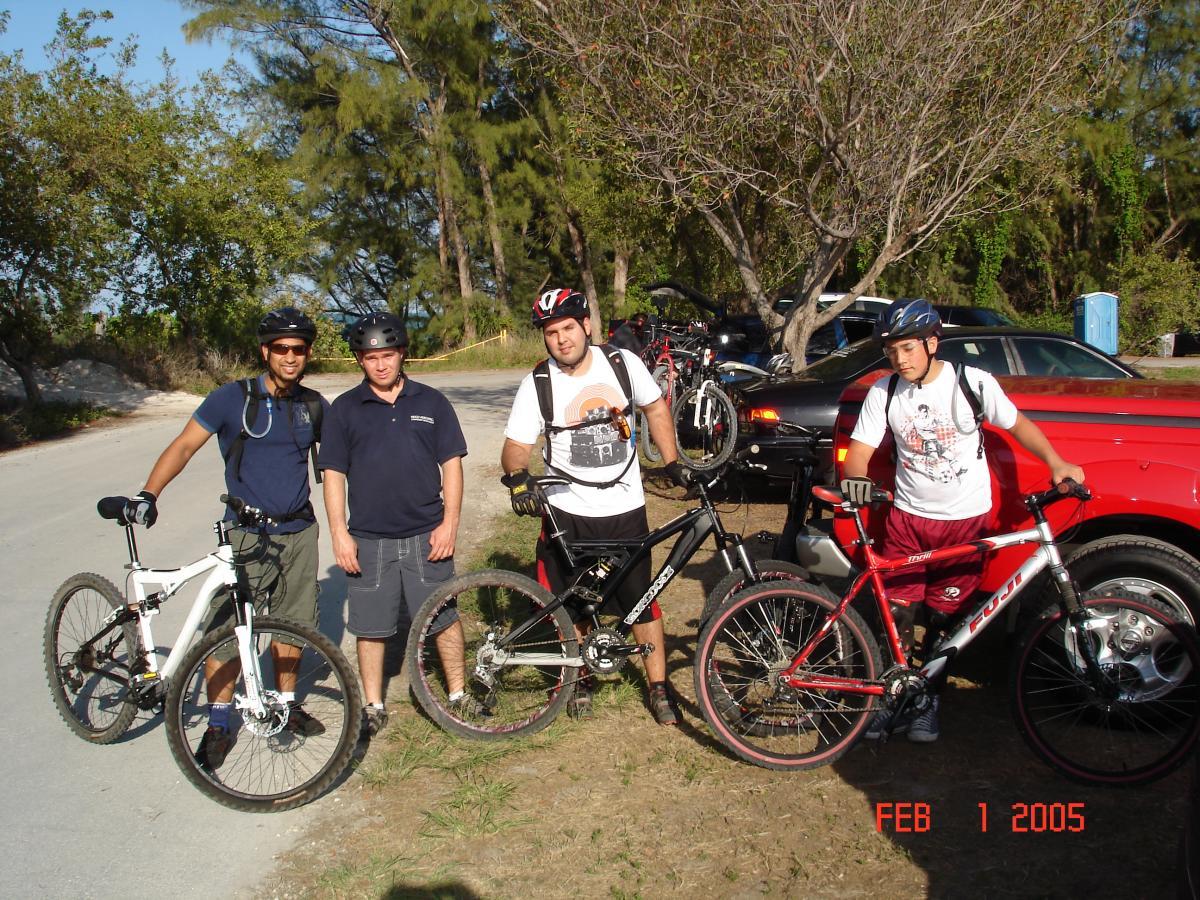 Four men are standing together near their bicycles, which are parked on a grassy area beside a road. Two of the men are wearing helmets and casual cycling attire, while one is dressed in a short-sleeved shirt and shorts. They appear to be enjoying a day of mountain biking. In the background, there are trees and a pickup truck with several bicycles mounted in the bed. The date in the corner of the image reads February 1, 2005. Virginia Key North Point mountain bike trail.