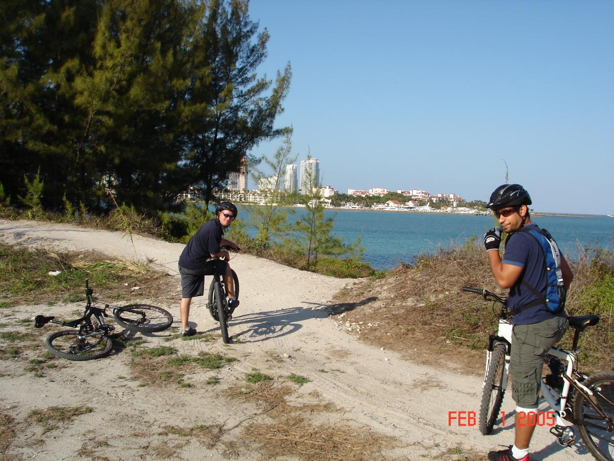 Two cyclists are on a dirt path near a body of water, surrounded by greenery. One cyclist is bending down to fix a bike, while the other is standing beside their bike, smiling and giving a thumbs up. In the background, there are buildings visible along the shoreline under a clear blue sky. Virginia Key North Point mountain bike trail.