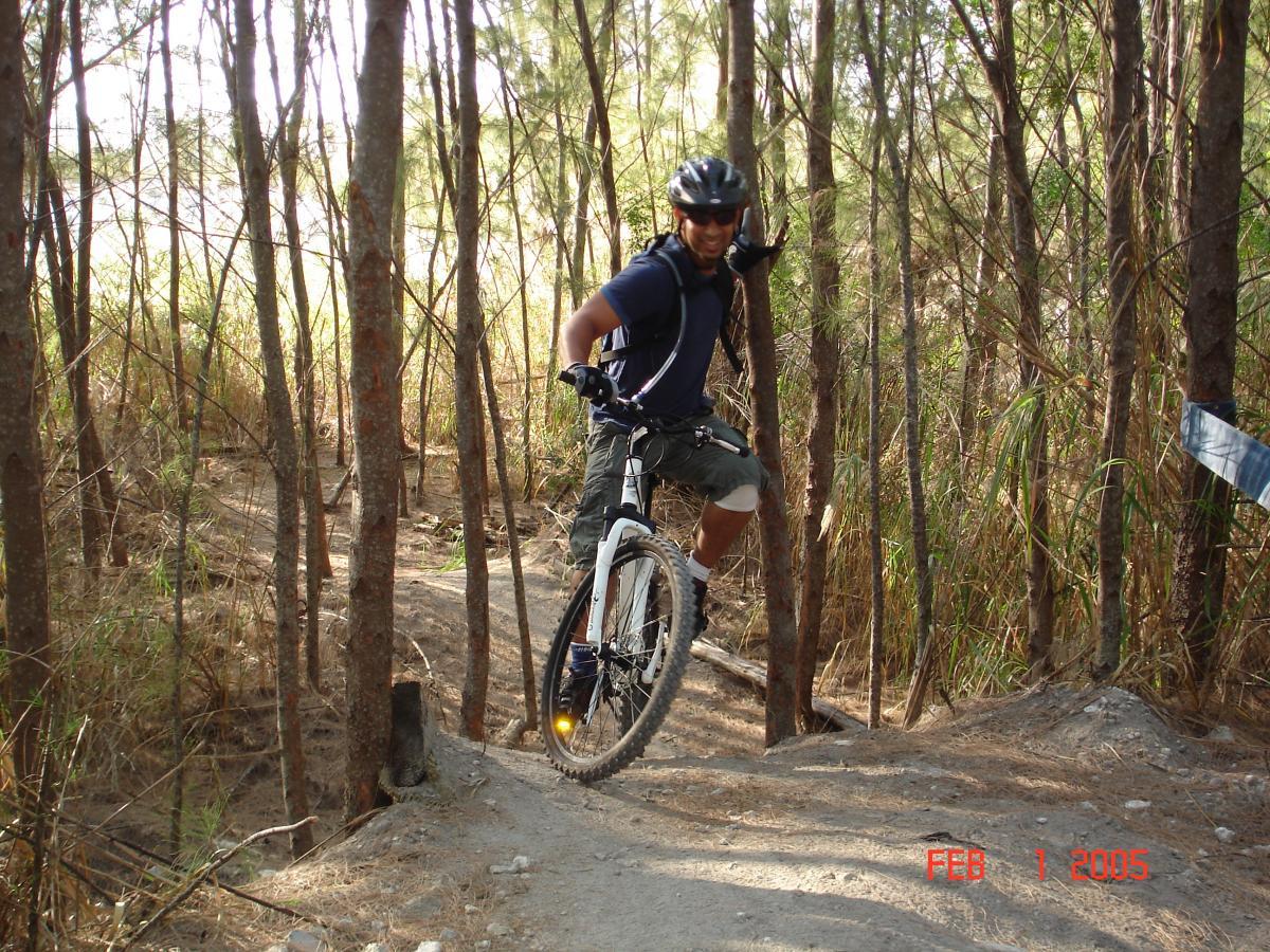 A person wearing a helmet and cycling gear is posing beside a mountain bike on a dirt trail surrounded by trees. The individual is smiling and giving a thumbs-up, with the sunlight filtering through the foliage in the background. The scene captures the spirit of outdoor adventure and mountain biking. Virginia Key North Point mountain bike trail.