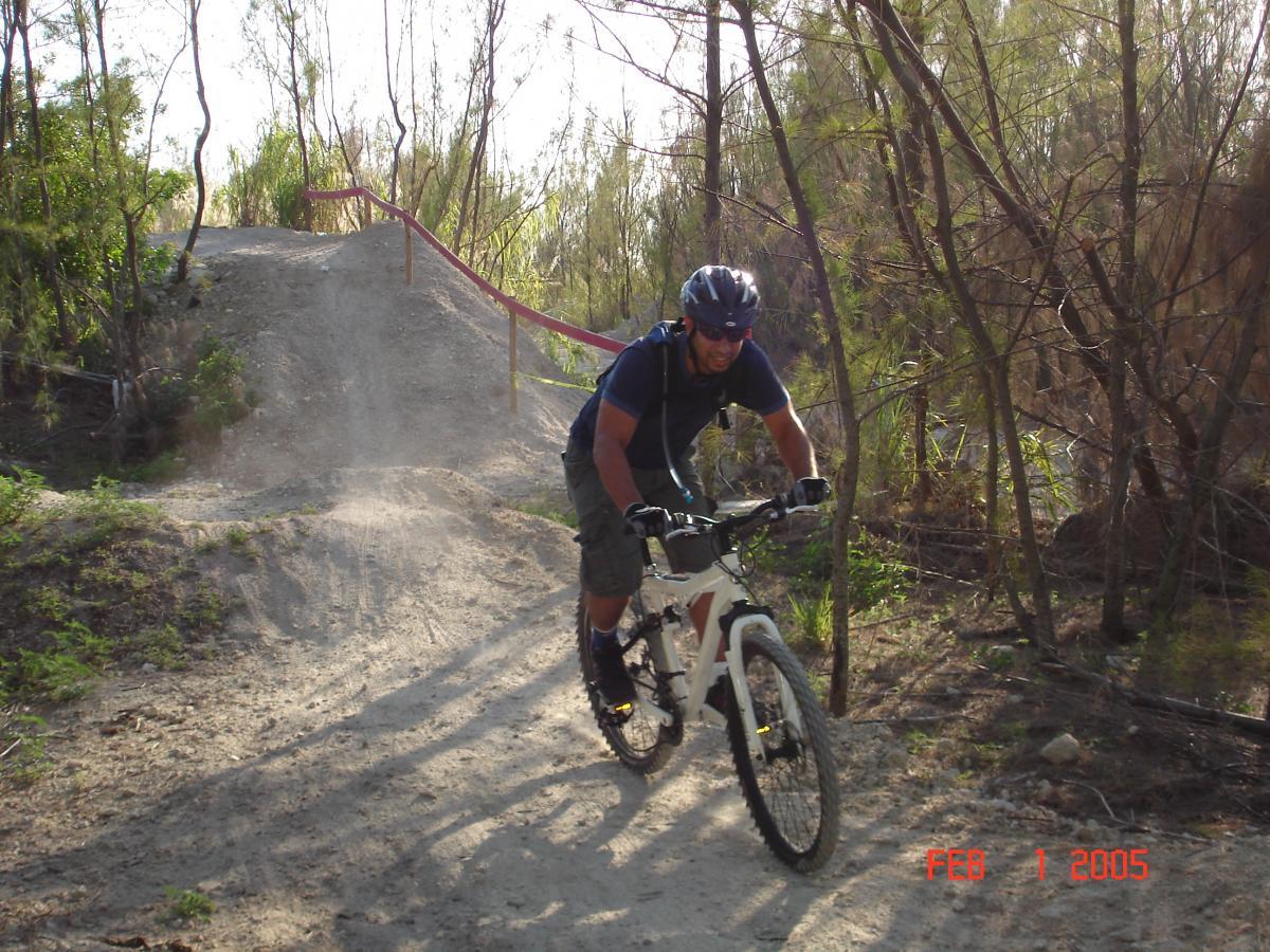 A mountain biker riding a white bicycle down a dirt trail, with dust kicking up behind him. He is wearing a blue helmet and a blue shirt, surrounded by trees and greenery. The sun is shining, creating a bright day for outdoor cycling. Virginia Key North Point mountain bike trail.