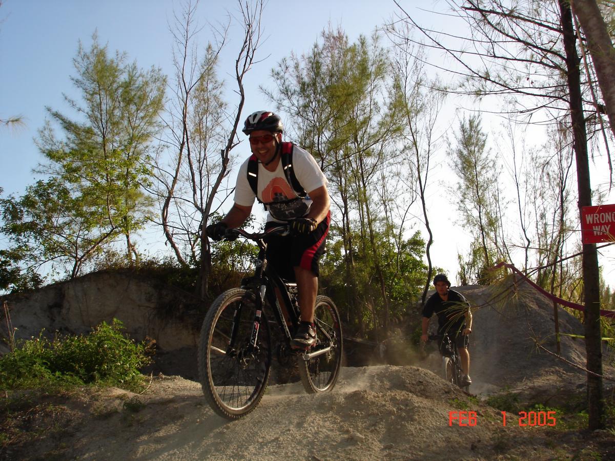 Two mountain bikers navigating a dusty trail surrounded by trees on a sunny day. The rider in the foreground is smiling and wearing a helmet and casual biking attire, while the cyclist in the background is also in biking gear. A "Wrong Way" sign is visible in the background. Virginia Key North Point mountain bike trail.
