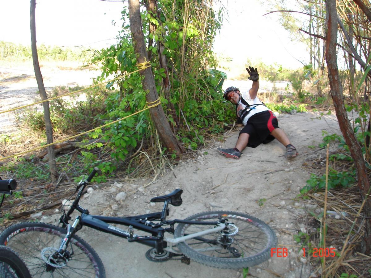 A person wearing a helmet and gloves is sitting on the ground next to a mountain bike, smiling and waving. The scene features a sandy trail surrounded by greenery, with a yellow rope tied to nearby trees. The image is dated February 1, 2005. Virginia Key North Point mountain bike trail.
